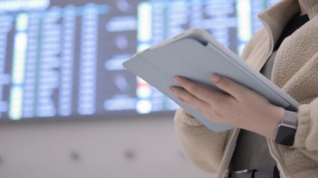 Person holds tablet near airport departure board. Blurred background shows flight information screens. Person wears beige coat and smartwatch. Focus on hands and device. Airport setting with travelers