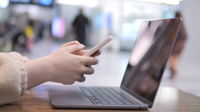 Person uses laptop and phone in public space. Hands type on keyboard, hold phone. Background shows blurred people and lights. Scene captures modern multitasking behavior. Focus stays on hands