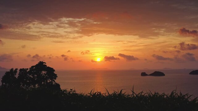 Beautiful orange sunset over the ocean near Koh Samui, Thailand, with silhouetted islands in the distance and tall grass foreground, creating a tranquil tropical scene during dusk. Nature background