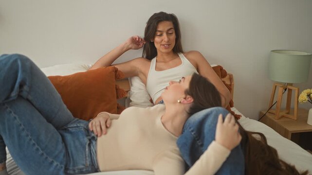 Two women on a bed in a studio bedroom set, one resting head on the other's lap while she smiles and tucks hair behind ear; comfort.