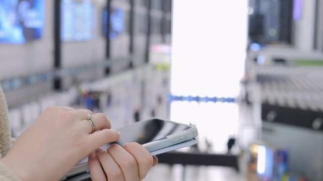 Close-up of hands holding and using a tablet in a modern transport hub. Background features blurred digital screens and bright lighting. Person wears a light-colored sweater and has rings on fingers