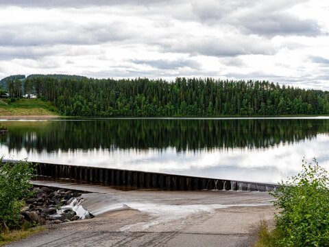Calm reservoir and overflow dam at Spegeldammen in Vuollerim, northern Sweden, reflecting a dense boreal forest beneath a dramatic cloudy sky. A peaceful Scandinavian landscape showcasing the natural 