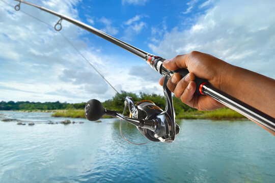 Closeup of hand holding fishing rod and spinning reel over lake under blue sky