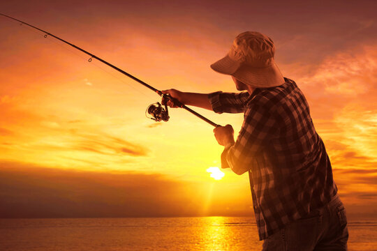 Back view of man wearing hat fishing by ocean at sunset