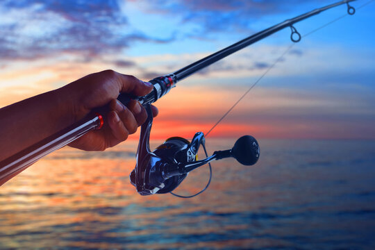 Closeup of hand holding fishing rod and spinning reel at ocean with sunset sky