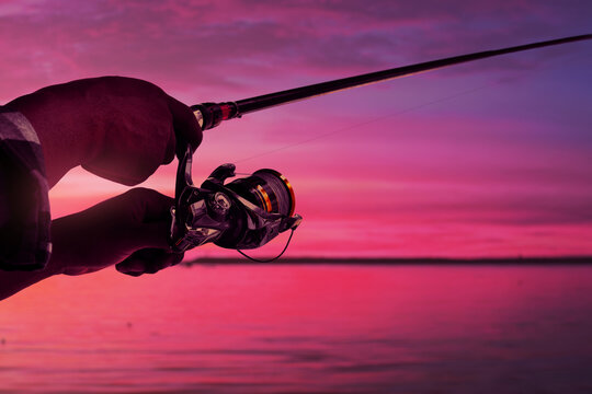 Closeup of hand holding fishing rod and spinning reel at ocean with sunset sky