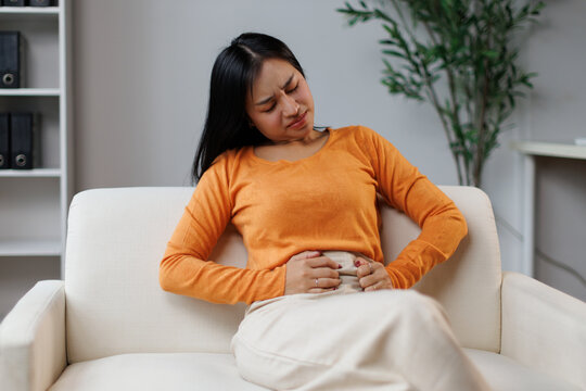 Young woman sitting on a comfortable white sofa. Experiencing intense abdominal pain and discomfort. Holding her stomach and bending over with a pained expression