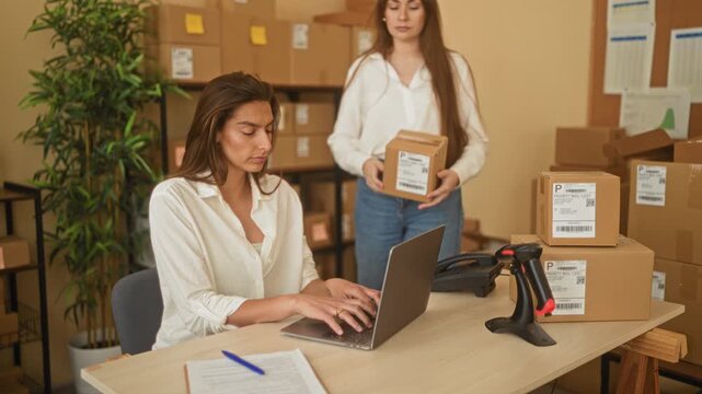 Woman points finger to parcel label while colleague holds a shipping package at a small business office fulfillment desk in a building; teamwork focus efficiency.