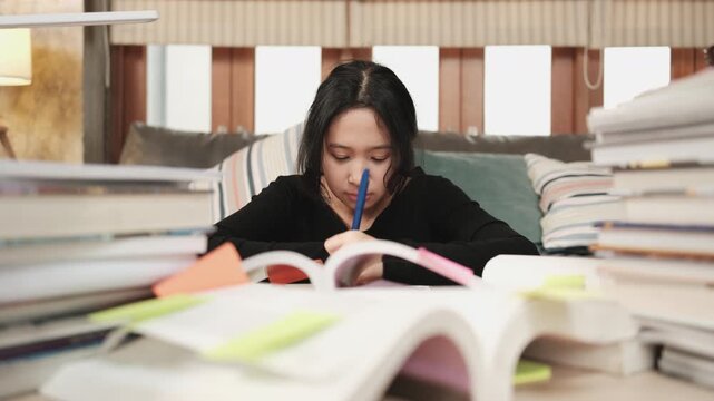 Asian teenage girl concentrates on pile of books, focuses on learning at home with stack of textbooks, reading and writing, self-study for school, Homeschooling, university education lifestyle.