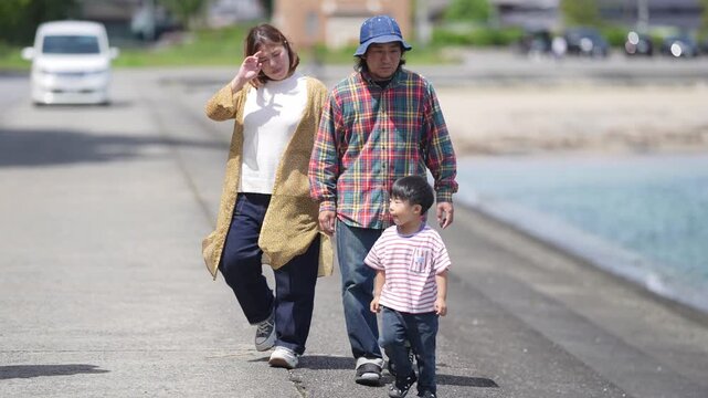 Sunlit family walks seaside path. Mother shields eyes from sun. Grandfather wears blue cap. Child runs beside grandparents. Grandparent holds infant gently. Scene evokes peaceful, warm day