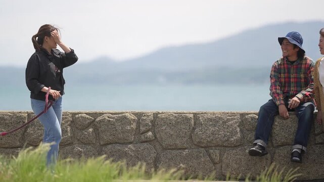 Woman walks with dog near sea. Man sits quietly on stone wall. Both overlook calm waters under soft sky. Cozy seaside moment, gentle, peaceful. Suitable for travel, nature, or quiet lifestyle scenes