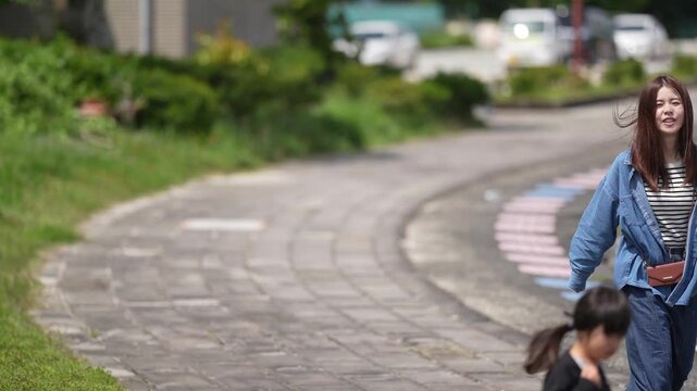 Mother smiles walking with two young children. Children run ahead on a paved path. Background shows greenery and parked cars. Footage captures joyful family moments outdoors