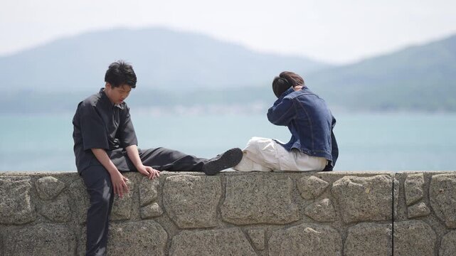 Two men sit alone on a stone wall. One rests head in hands, shoulders slumped. The other leans forward, lost in thought. Distant hills and calm sea frame their quiet moment