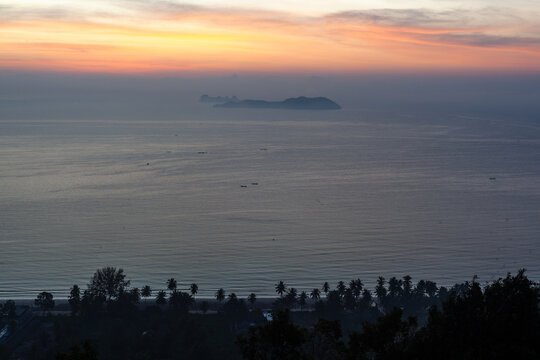 Khao Phang Pang Viewpoint at sunset. Calm ocean waters with distant island silhouette. Palm trees lining the shore. Chumphon, Thailand coastal scenery.