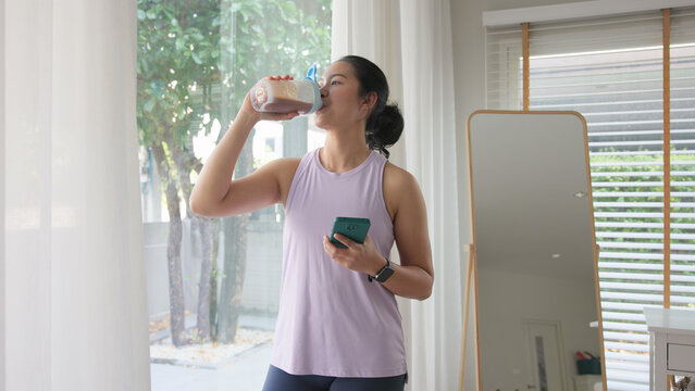 Young gen z active woman drink whey creatine shake bottle eating diet food using phone calorie plan app after home sport workout. health care asia people happy relax protect aging life weight loss.