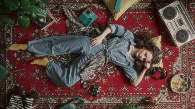 Directly above view of young curly-haired woman in denim jumpsuit and yellow socks lying on red patterned carpet next to vintage stereo, rotary phone and old camera. Video portrait