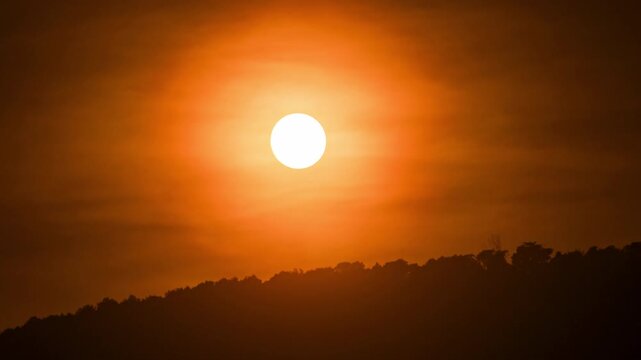 Time-lapse of the sun disappearing behind dark mountain silhouettes under a majestic sky.