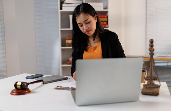 Asian businesswoman or female lawyer working on legal paperwork in a modern law office, using a laptop and writing documents, surrounded by justice symbols