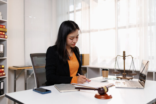 Young professional woman reviewing legal documents, using a pen at her office desk with a gavel and scales of justice, symbolizing law, legal counsel, and business support