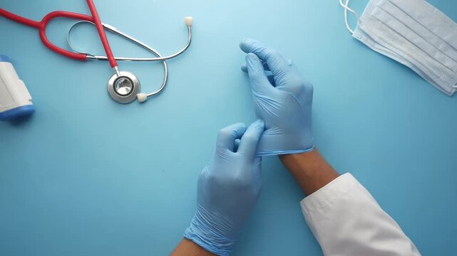 Close up of doctor hands putting on blue protective medical gloves with stethoscope and mask on blue background