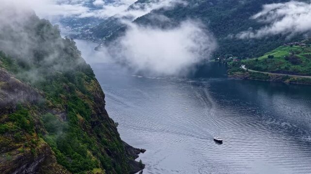 Aerial timelapse of vessel moving through Norway fjord, dramatic mountains, mist, scenic landscape.