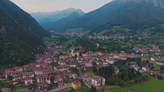 Aerial drone forward tracking shot flying over Bezzecca village rooftops with yellow church toward deep alpine valley, layered mountain ridges in background