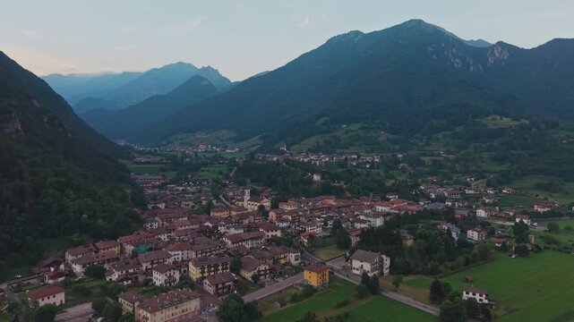 Aerial drone pullback shot revealing Bezzecca village with terracotta rooftops and church bell tower surrounded by green meadows, layered mountain ridges in background