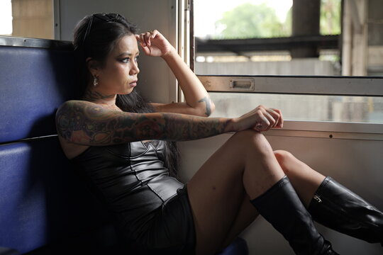 Young heavily tattooed Asian woman wearing a black leather dress and knee-high boots sits pensively inside a moving train, gazing out the window with sunglasses resting on her head