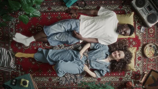Directly above view of young Black man and Caucasian woman chatting and smiling while relaxing on vintage patterned rug, surrounded by 90s analog electronics and retro items