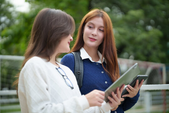 Two female college students use a tablet and smartphone while talking outdoors on campus, representing friendship, digital learning, collaboration, and modern education.