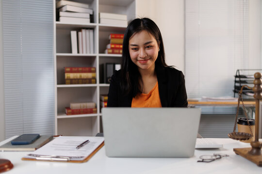 Young asian woman lawyer or attorney is concentrating on her work, using a laptop at a bright office desk with legal documents and a scale representing justice
