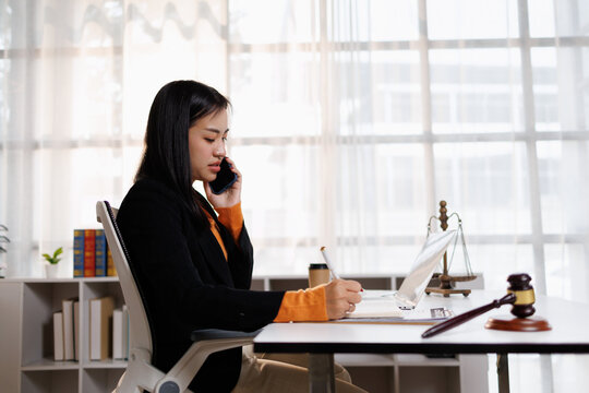 Young asian woman lawyer managing her legal work, talking on a mobile phone and writing important details in a notebook, symbolizing dedicated professional service and communication