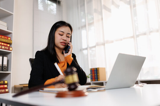Young asian female lawyer discussing legal case on smartphone, writing notes at office desk with gavel and laptop, representing justice and law profession