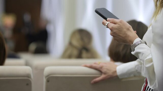Woman holds phone while seated in modern auditorium. She glances at screen, perhaps checking messages or notes. Her calm posture suggests focus during event. Background shows blurred audience