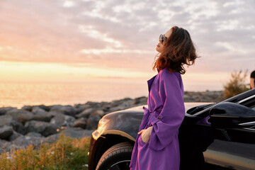 Fototapeta premium A woman in a purple coat stands beside a sleek car at sunset, gazing into the distance. A lifestyle moment in the outdoors blends fashion, travel and calm confidence.
