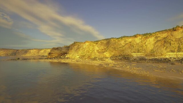 textured golden cliffs basking in warm light, rugged rockface and visible strata create dramatic contrast, scattered pebbles at base, long shadows and bold