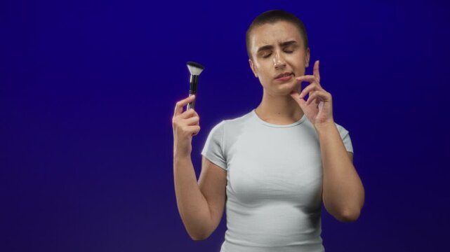 Woman holding makeup brush with finger on chin in studio, short buzzed hair, white tee, examining brush near face against deep blue backdrop; uncertainty.