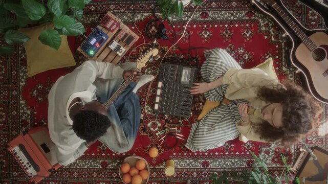 Top view of two diverse creative musicians sitting on cozy red rug, man playing electric guitar and woman singing into microphone while recording audio with retro cassette tape multitrack recorder