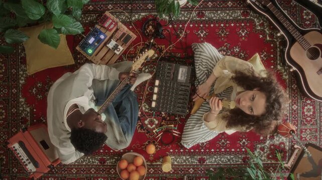 Overhead view of creative duo sitting on red patterned rug among retro recording devices, Black man playing guitar while Caucasian female musician looking up at camera and singing into microphone