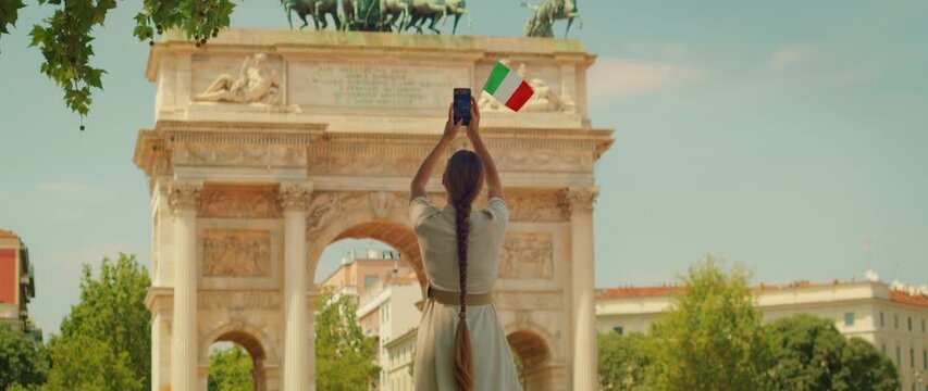 Back view of tourist raising phone and capturing photo of historic arch in Milan