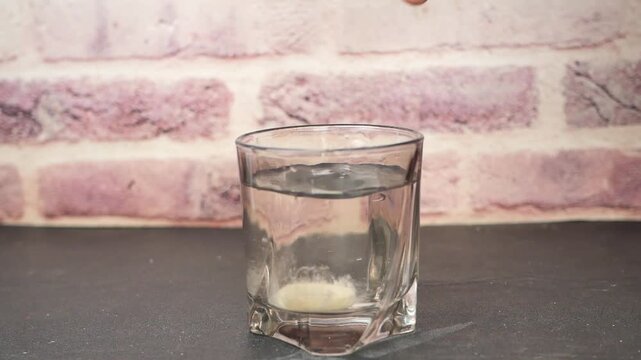 Close up slow motion video of hand dropping an effervescent tablet into glass of fresh clear water on dark table with brick wall background