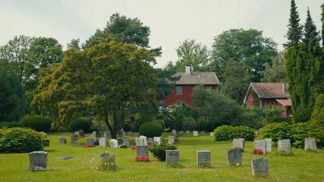 A quiet graveyard in Oslo, Norway stretches across a green lawn beneath mature trees, with red wooden houses in the background adding a distinctly Scandinavian sense of place and everyday stillness.