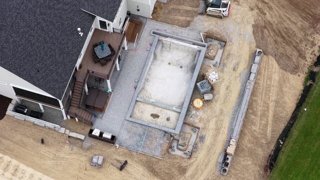 Empty pool shell sits beside wooden deck with hot tub in suburban backyard under construction in USA, dirt covers graded yard near delivery truck plus staging materials. Aerial drone top down