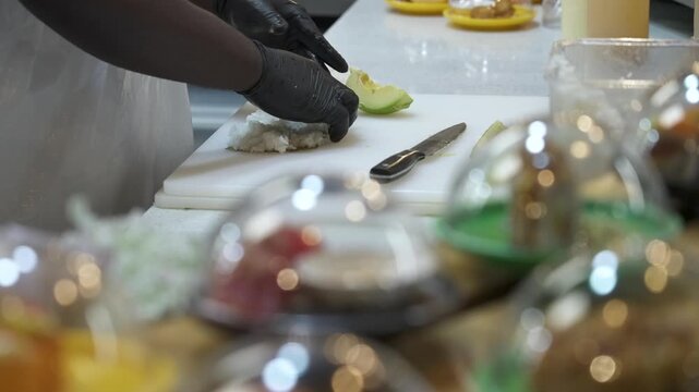 Food worker in black gloves presses rice on a cutting board while prepared sushi plates move past in the foreground.