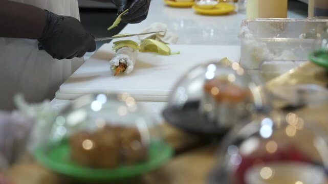 Gloved hands cut an avocado covered roll on a cutting board as plated items pass on a conveyor at an in-store sushi restaurant.
