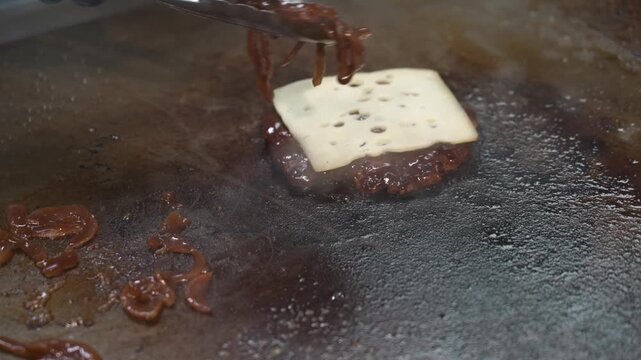 Cook uses tongs to place caramelised onions onto a cheese-topped burger patty sizzling on a flat top grill.