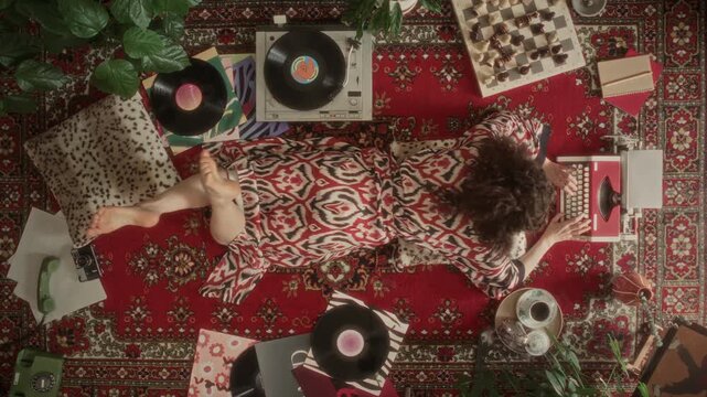 High angle shot of curly-haired woman in robe lying prone on patterned rug with her feet raised, typing on vintage typewriter and enjoying music from retro vinyl record player