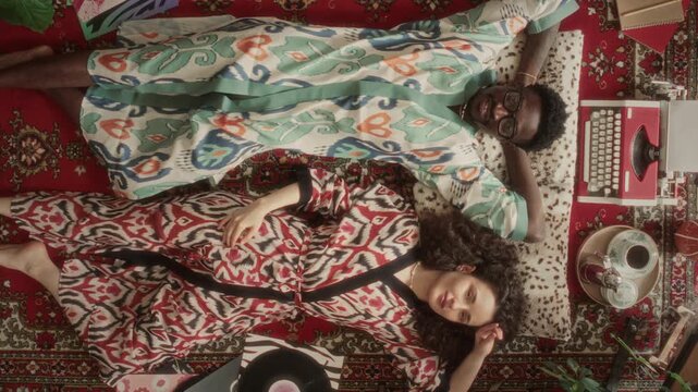Directly above view of smiling young multiethnic couple in patterned robes, lying down on red rug and looking at camera, surrounded by retro vinyl records and typewriter. Video portrait