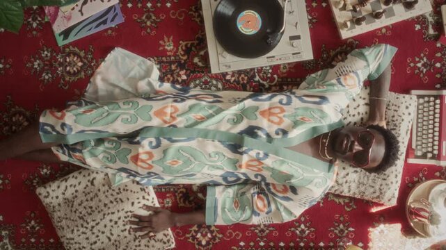 High angle portrait of young African American man in patterned robe and sunglasses relaxing with one hand behind his head and enjoying vinyl music from analog turntable on retro-style red carpet