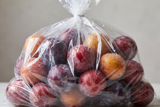 Ripe red plums packed inside a clear tied plastic bag sitting on a bright surface